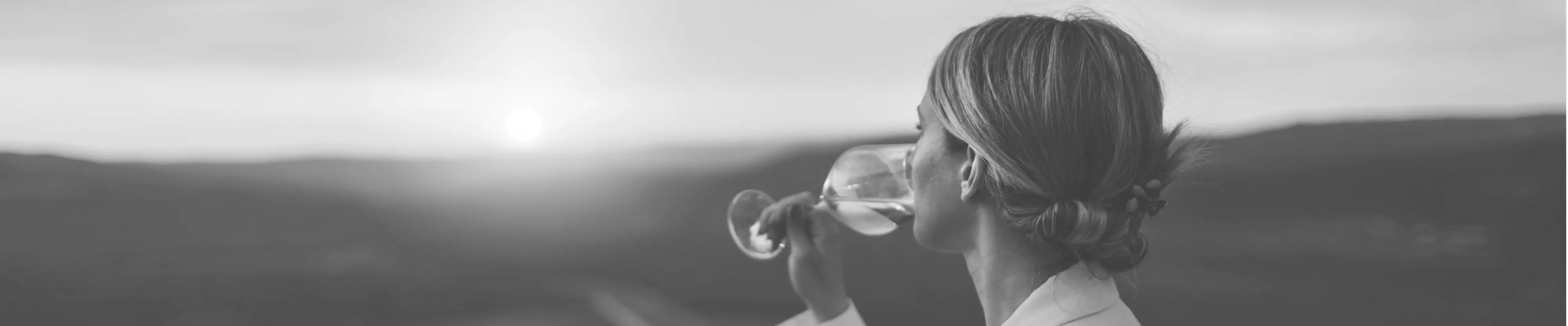 Schwarz-Weiss Bild von Frau, die in die Ferne schaut und ein Schluck Wein aus einem Glas trinkt.